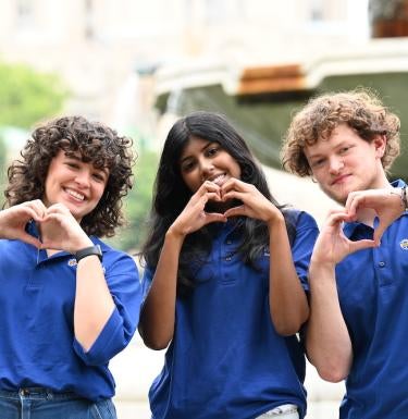 Three students make heart shapes with their hands in front of a fountain