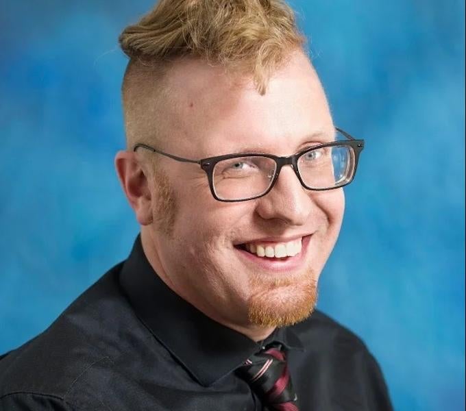 Andrew Lotz, man with red hair and thick-framed glasses against blue backdrop